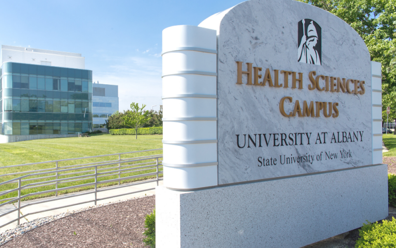 A marble sign says "Health Sciences Campus". Behind the sign, green grass and a blue sky frame the Cancer Research Center.
