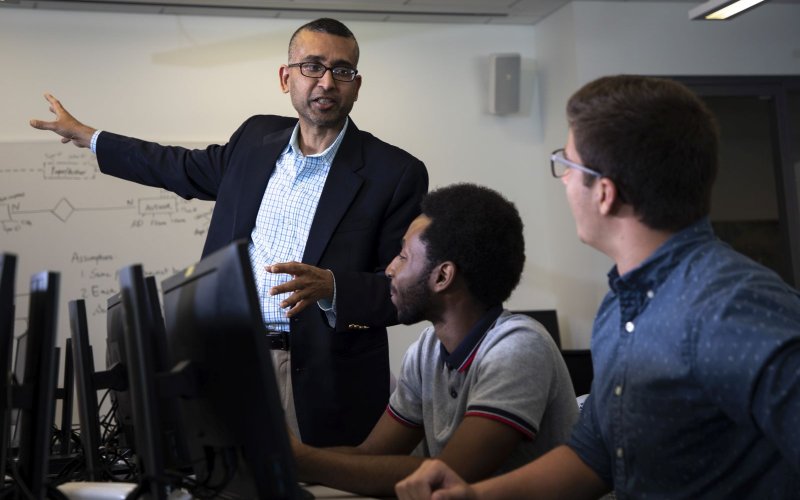 Professor Sanjay Goal gestures toward a whiteboard covered with diagrams while talking to two students seated at a table filled with computers.