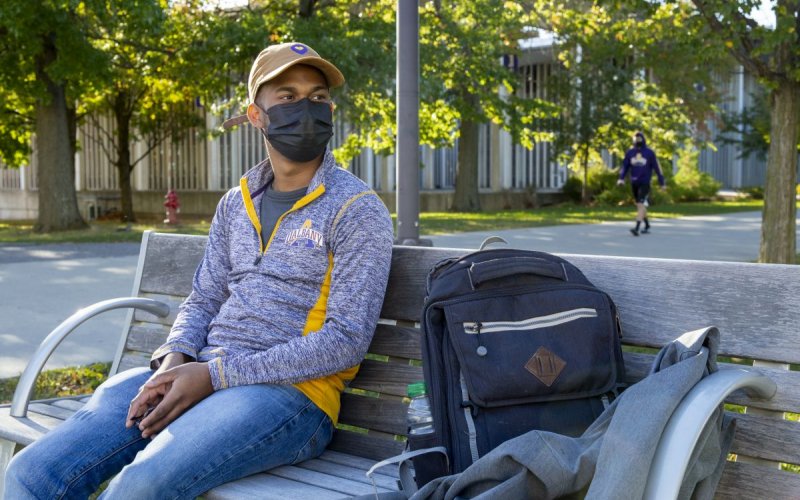 A student in a baseball cap and face mask sits on a park bench with his backpack