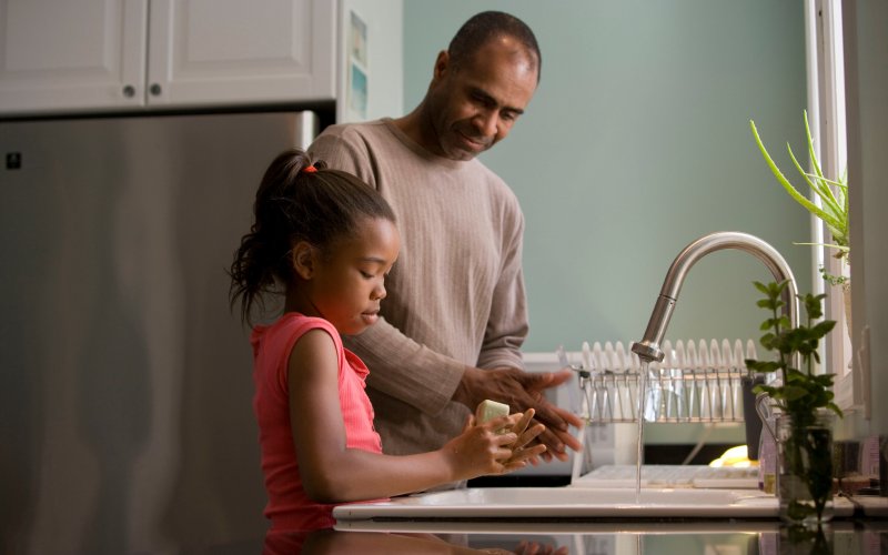 A man and his daughter stand at a sink, washing their hands together.