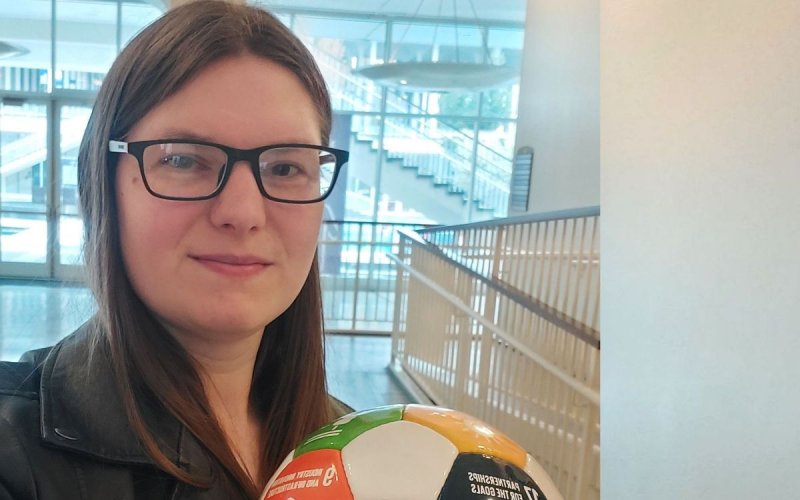a young woman with brown hair and glasses holds a soccer ball imprinted with the UN Sustainable Goals
