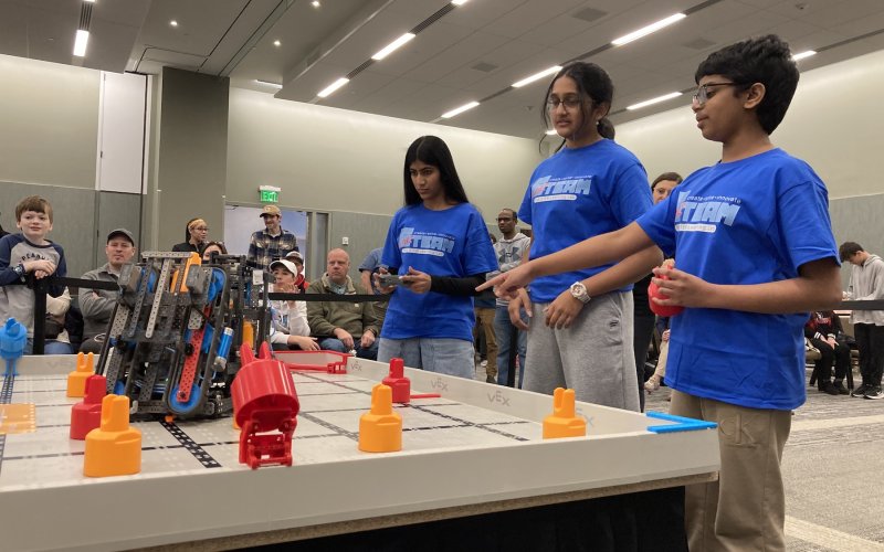 Two girls and a boy, all in blue tee shirts, stare and point at a robotics game board 
