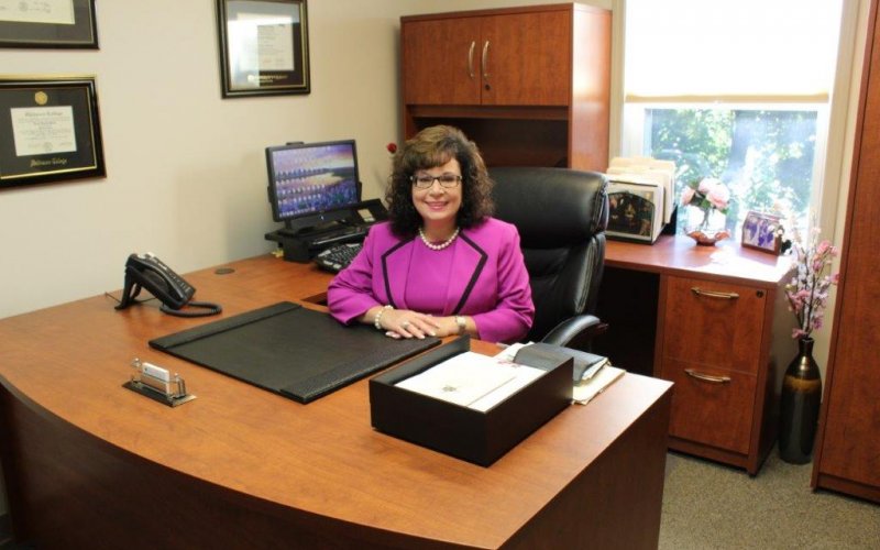 Dr. Nicole MacFarland, wearing a hot pink suit, sits behind her wooden desk in her office at Senior Hope Counseling. She is smiling at the camera.