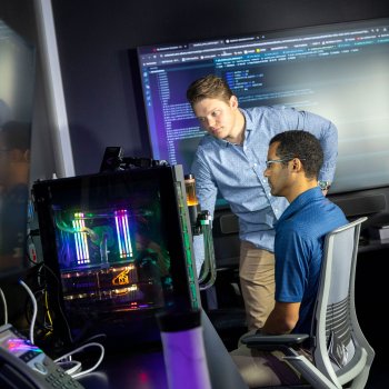 One researcher sits in a chair in front of several computer monitors while a second researcher leans over and says something while looking at the computer.
