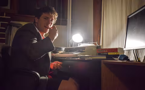 A young man sits at a desk in a dark room, typewriter before him, illuminated by one small desk lamp.