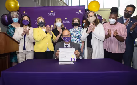 President Rodríguez sitting at a table and holding up the signed Okanagan Charter, surrounded by members of the Well-being Collective and members of the University administration.
