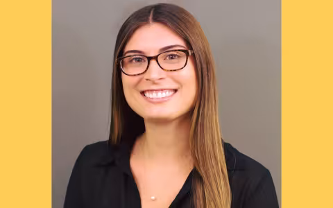 Sebesta shown smiling in a black shirt with glasses on, yellow border around photo.