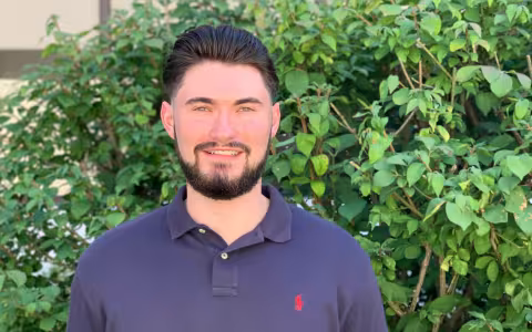 CDPHP Scholar, CJ Gil, outside in front of greenery. He has dark hair, facial hair, and is wearing a navy blue polo.