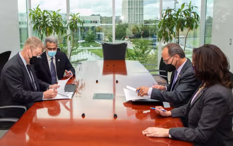 Four people in facemasks and business attire sign papers at a long wooden table in front of a large window with house plants in front of it.
