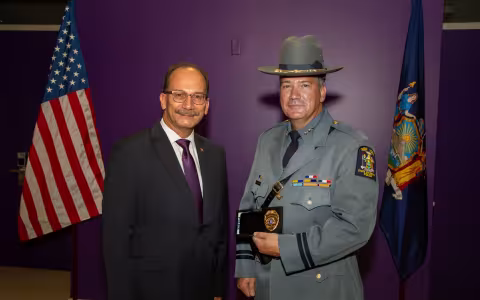A smiling man in a dark suit and an officer in State Police uniform stand between the U.S. and N.Y. flags