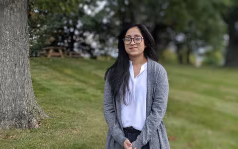 Thoin Begum, a PhD student, stands outside in a field, looking at the camera. Picnic tables and trees are in the background.