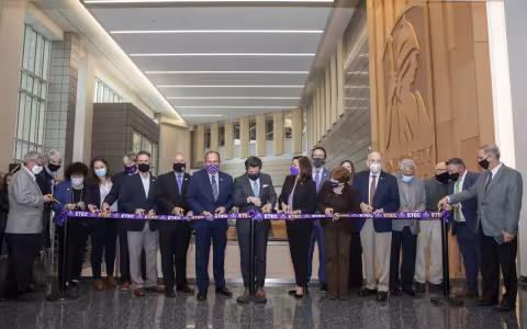 18 people in business clothes stand with scissors, ready to cut a purple ribbon stretched in front of them with the word ETEC repeated on it