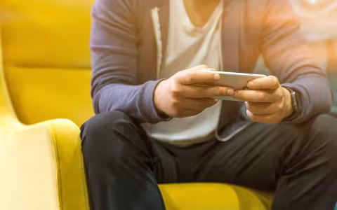 A person, pictured only below the shoulders, sits on a yellow chair while looking at their phone.
