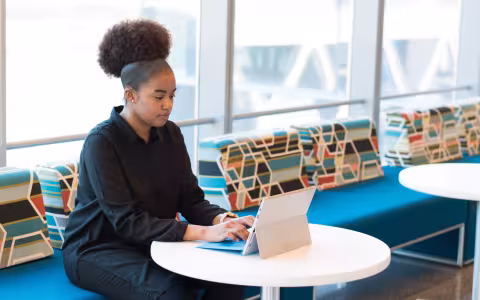 A woman sits at a table, looking at a white tablet propped up next to a keyboard.