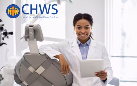 A female doctor leans against a dentistry chair and holds a tablet.