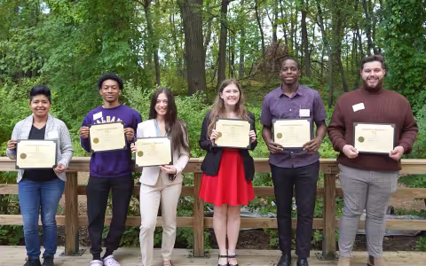 Six smiling students stand in a row in front of a wooden fence, each holding a scholarship certificate