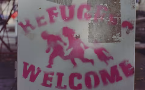 a sign stencilled in pink spray paint shows three people running and the word "refugees welcome"