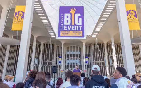 A man speaks to a group of students and staff from an outdoor podium in front of the campus center. A hanging banner reads UAlbany's Big Event.