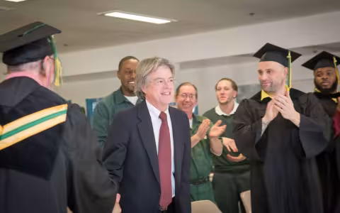 Randal Craig, smiling in a blue suit and red tie, is surrounded by clapping men in graduation garb or prison garb