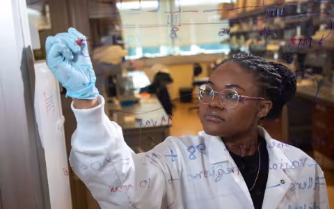 A woman in a lab coat and blue goves is seen behind a pane of glass she is writing formulas on.