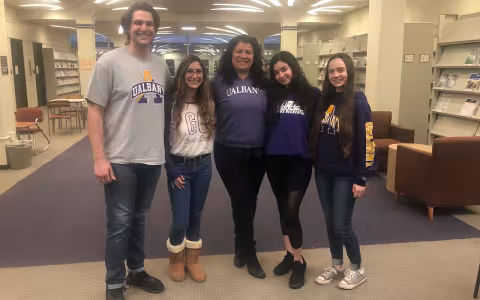 A tale male student and 3 female students and female professor stand in line in a library conference room
