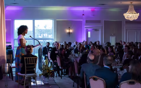 A woman in a long skirt speaks at a podium to a crowd seated at dinner tables