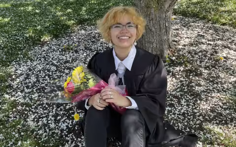 Maya Lee '22 in glasses holds a bouquet of flowers and offers a big smile while sitting under a tree