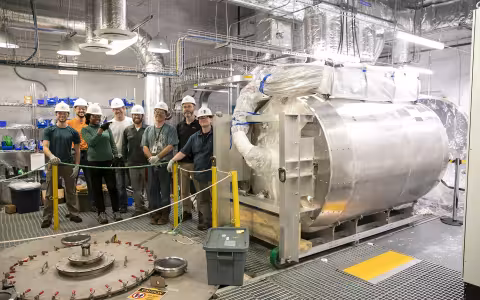 A group of scientists and movers stand near a large cylinder dark matter tank inside of a lab room.