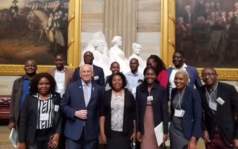 A group of smiling people pose inside the U.S. Capital in front of a sculpture and two paintings