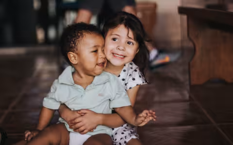A small child is sitting on the floor, hugging a toddler who sits on her lap. 