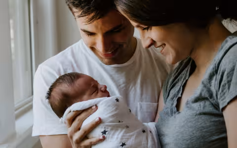 A man and a woman are holding a baby swaddled in a blanket and are smiling at the baby.
