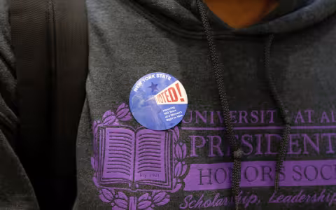 A round "New York State VOTED" sticker is seen on a student's sweatshirt, which partially shows the words "Univerisity at Albany Presidential Honot Society: Scholarship, Leadership"