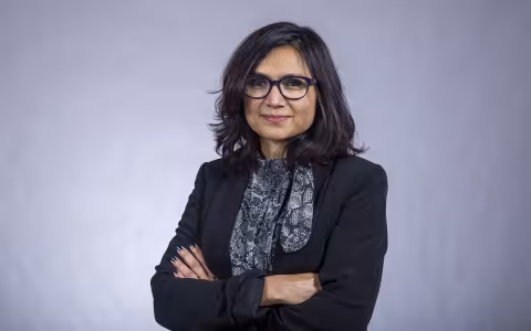 A portrait of Associate Professor Rukhsana Ahmed smiling with arms crossed in front of a plain gray background.