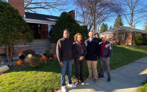 Four people in jackets stand on a sidewalk outside a brick house with a lawn decorated pumpkins.