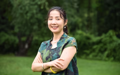 Youqin Huang, arms folded, smiles and poses for a photo while standing outdoors.