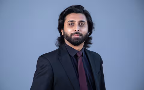 Portrait of a man with shoulder-length black hair and black beard, wearing a black suit jacket and purple tie, against a blue background.