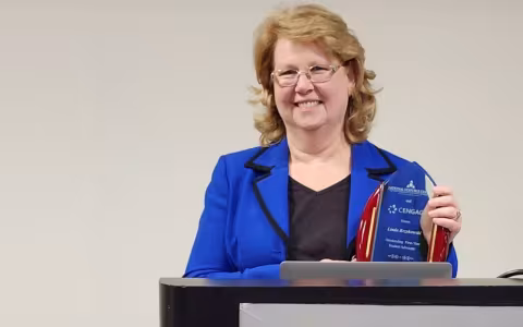 A woman with shoulder-length blonde hair and glasses in a blue blazer holds an award at a podium.