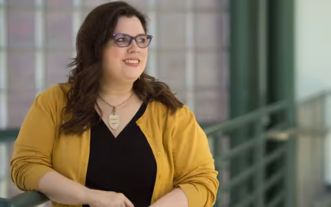 Dr. Melanie Sage wears a black top with yellow sweater, a gold necklace and purple rimmed glasses, leaning against a green railing indoors, smiling as she looks off to the side