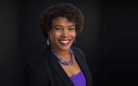 A smiling woman with curly brown hair, silver earings and a beaded necklace