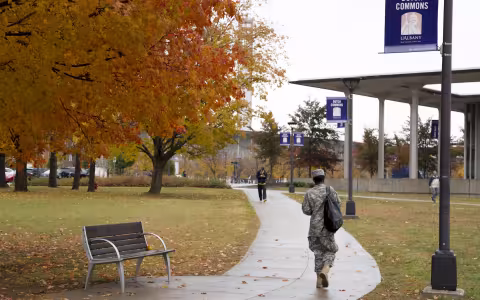A person in a military uniform can be seen from behind walking along a path through Dutch Commons on the UAlbany campus on a wet fall day. A student walks toward them and trees display bright orange and yellow leaves.