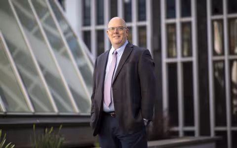 A man in a suit jacket and tie stands near glass windows