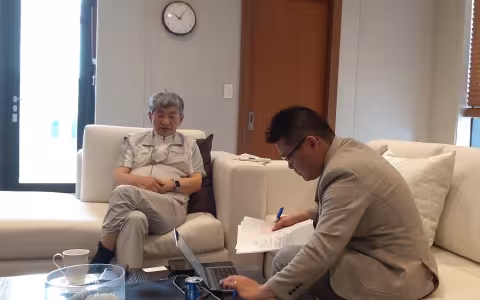 An older man with silver hair is seated in a chair next to a younger man with black hair and glasses, who sits on a couch nearby consulting a handful of papers and looking at a laptop. They are gathered around a table with a pitcher of water and other beverages.