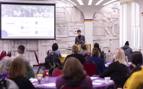 A woman speaks from a podium near a screen with a projected agenda on it. The backs of heads of the audience, seated at round tables, is seen in the foreground.