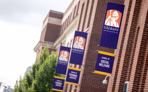 UAlbany's School of Social Welfare building on the Downtown Campus. The building is red brick, with three sets of purple and gold UAlbany flags mounted on the side. Each set of flags has the Minerva logo on top. Then below, one flag says School of Social Welfare; the other two say School of Criminal Justice.