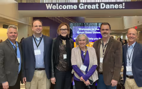 Six people in business attire and lanyards pose in the UAlbany Student Union. Behind them is a digital sign that reads "Welcome to the Great Dane Union", and "Welcome Great Danes" is painted on the wall above.