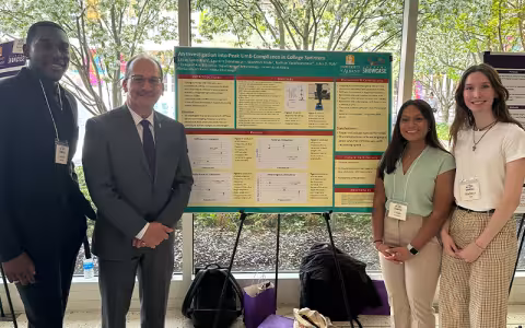 Students, with UAlbany President Havidán Rodríguez stand on either side of a presentation board titled, "An Investigation into Peak Limb Compliance in College Sprinters." President Rodríguez is wearing a gray suit and the students are dressed professionally and wearing lanyards.