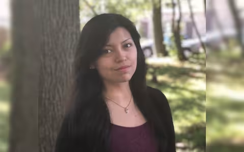 A woman with dark hair, a maroon top and a black cardigan smiles at the camera. She stands in front of a tree. Grass, sunlight and a couple of cars are in the background.
