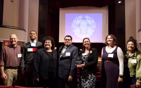 A group of seven people stand in an auditorium dressed in business attire. One holds an engraved award and the projector screen behind them reads, "Presidents Awards for Exemplary Public Engagement 2023."
