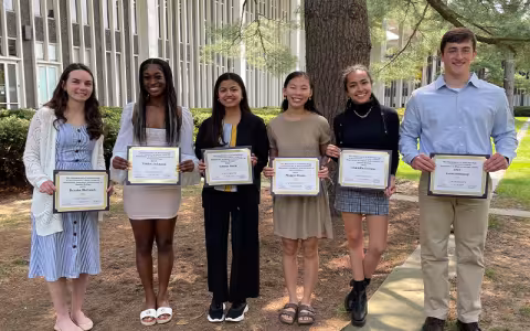 Six students stand outside on campus on a sunny day while holding award certificates.
