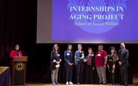 A woman stands at a podium near a line of seven smiling people standing under a screen with the words "Internships in Aging Project/ School of Social Welfare"